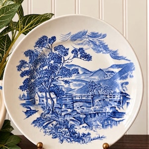 May include: A round ceramic plate featuring a blue and white transferware pattern of a rural landscape. The design depicts a stone bridge over a stream, a small cottage, large trees, and rolling hills under a sky with stylized clouds.