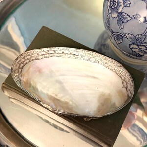 May include: An oval-shaped dish featuring a polished mother-of-pearl interior with iridescent white and cream tones. The rim is encased in a decorative silver-toned metal border with a repeating honeycomb or cobblestone pattern.