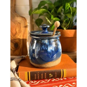 May include: A ceramic honey pot with a lid and a wooden dipper. The pot features a deep blue glaze that transitions into white drips over a natural tan base. It sits on a brown book titled Helen Corbitt's Cook Book with a mushroom icon.
