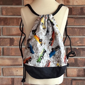 May include: A white drawstring bag with a black bottom. The bag is decorated with a pattern of vintage cars and road maps. The bag has black drawstrings.