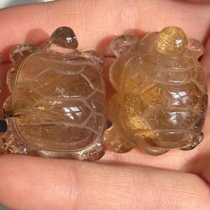 May include: Three translucent crystal turtle figurines in shades of amber and brown. Each turtle has a detailed shell pattern and is held in a hand. The turtles are small, decorative objects.