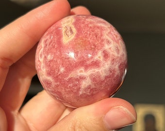 May include: A polished, spherical gemstone in shades of pink and white, held in a hand. The orb has a swirling, banded pattern and a glossy finish. The stone's colors range from deep rose to pale pink, with white veins.