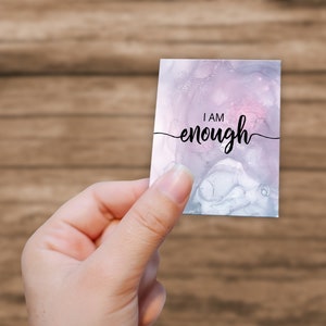 K&ouml;nnte beinhalten: Eine wei&szlig;e Karte mit einem rosa und grauen Aquarellhintergrund. Die Karte tr&auml;gt den Text "I AM enough" in schwarzer Kursivschrift.