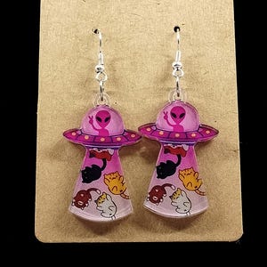 May include: A pair of silver dangle earrings featuring pink alien spaceships with cats inside. The cats are in various colors and poses.