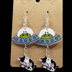 May include: A pair of dangle earrings featuring a cartoon cow and a UFO with a green alien inside. The cow is black and white with pink ears and is eating grass. The UFO is gray with a blue window and green accents. The earrings are silver.