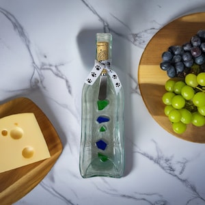 Puede incluir: Una botella de vidrio transparente con una cinta blanca y huellas de patas negras, utilizada como plato para servir queso y galletas. La botella tiene un pequeño cuchillo en su interior y está decorada con piezas de vidrio azul y verde.