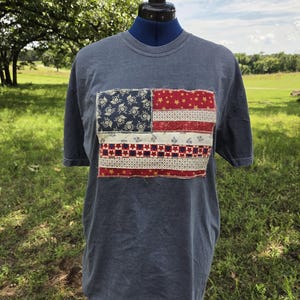 Può includere: Una t-shirt a maniche corte color blu ardesia con una toppa rettangolare sul petto. La toppa è disegnata per assomigliare alla bandiera americana, costruita con varie strisce di tessuto con motivi rossi, bianchi e blu, incluse stelle e fiori.