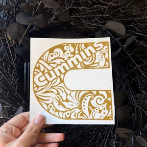 May include: A white square sticker featuring the word "Cummins" in a gold, ornate, stylized font. The letter "C" is the main design element, filled with floral and leaf patterns. The sticker is held against a dark background.
