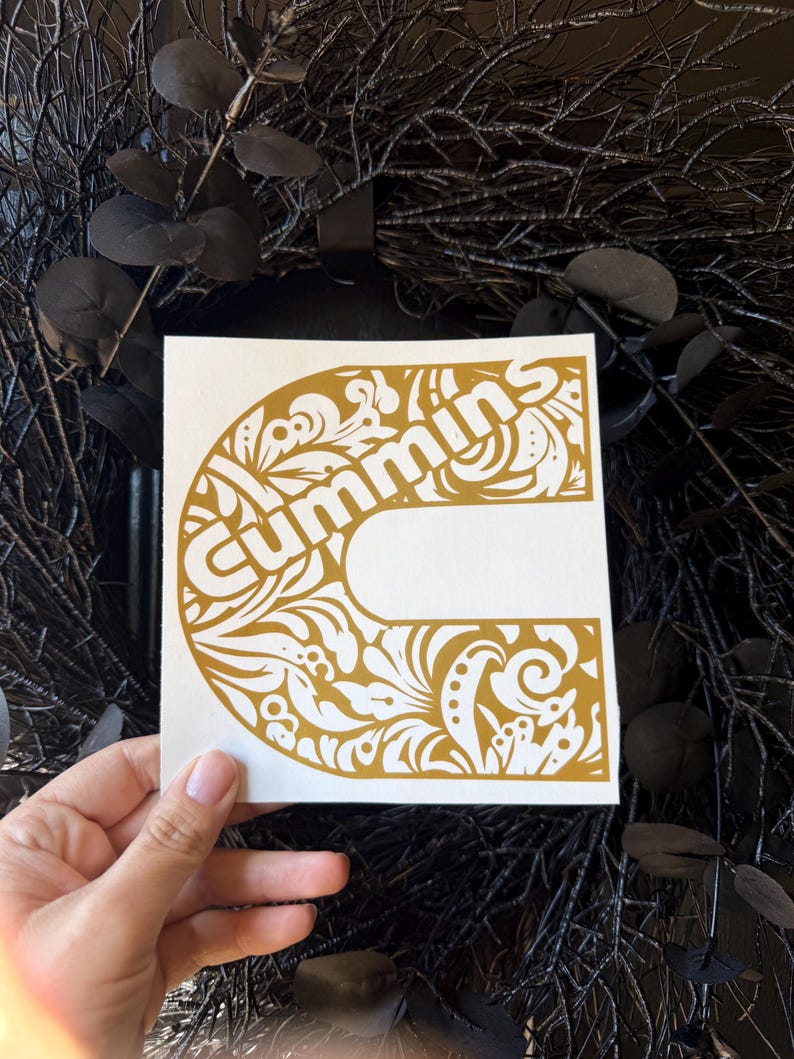 May include: A white square sticker featuring the word "Cummins" in a gold, ornate, stylized font. The letter "C" is the main design element, filled with floral and leaf patterns. The sticker is held against a dark background.