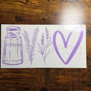 May include: A white decal featuring a purple salt shaker, sprigs of lavender, and a heart shape. The design is a simple, line-art style, perfect for home decor or crafting projects. The decal is on a wooden surface.