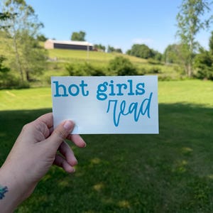 May include: A rectangular white vinyl decal featuring the text "hot girls read" in a bright cyan blue color. The text "hot" and "girls" are printed in a bold, lowercase serif font, while the word "read" is in a cursive script font.