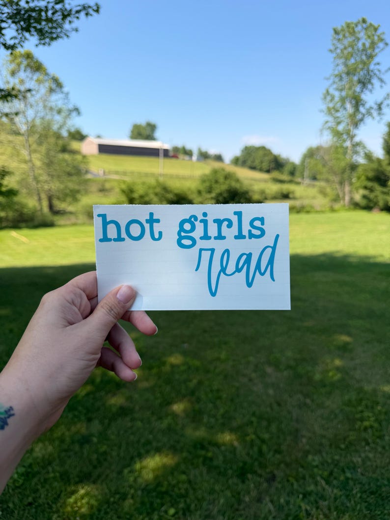 May include: A rectangular white vinyl decal featuring the text "hot girls read" in a bright cyan blue color. The text "hot" and "girls" are printed in a bold, lowercase serif font, while the word "read" is in a cursive script font.