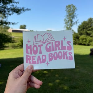 May include: A rectangular white sticker featuring the pink text "HOT GIRL'S READ BOOKS" in a bold, wavy font. Above the text is a simple line drawing of an open book, and small four-pointed star icons are positioned around the lettering.