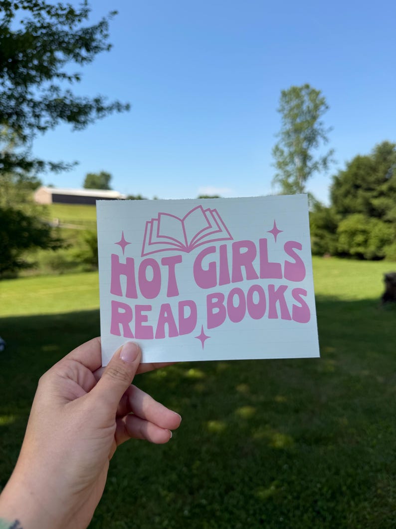 May include: A rectangular white sticker featuring the pink text "HOT GIRL'S READ BOOKS" in a bold, wavy font. Above the text is a simple line drawing of an open book, and small four-pointed star icons are positioned around the lettering.