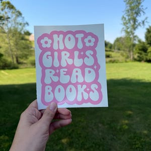May include: A square white sticker featuring the pink text HOT GIRLS READ BOOKS in a wavy, retro-inspired font. The design is framed by a pink scalloped border with small flower icons at the top corners and four-pointed star accents throughout.