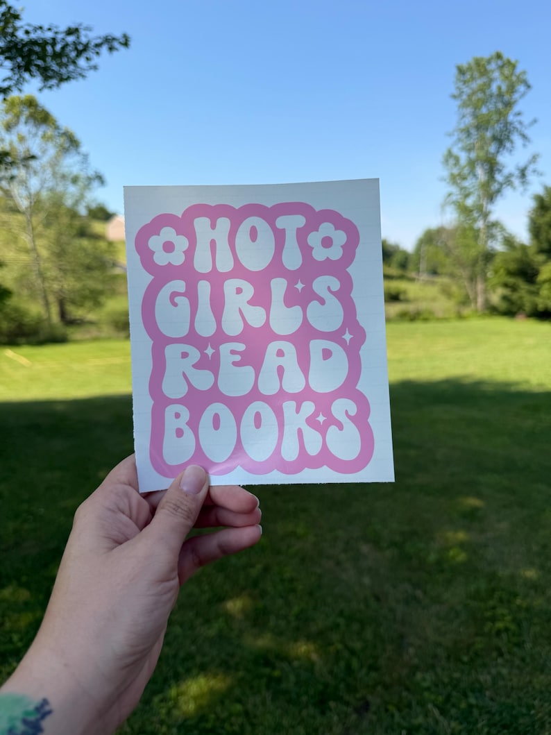 May include: A square white sticker featuring the pink text HOT GIRLS READ BOOKS in a wavy, retro-inspired font. The design is framed by a pink scalloped border with small flower icons at the top corners and four-pointed star accents throughout.