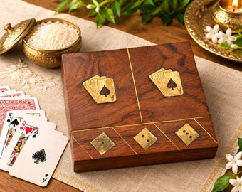 May include: A square wooden box featuring brass inlays of playing card suits and pips on the lid. The box is accompanied by a fanned-out deck of playing cards, a small brass bowl filled with white rice, and a brass candle holder with a lit candle.