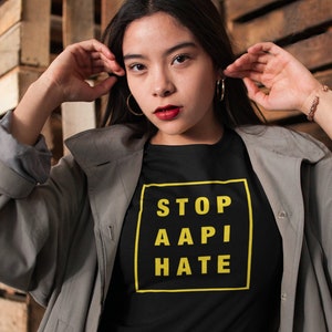 May include: A person wearing a black t-shirt with a yellow box that says "STOP AAPI HATE" and a gray jacket. The person is standing in front of a wooden wall.