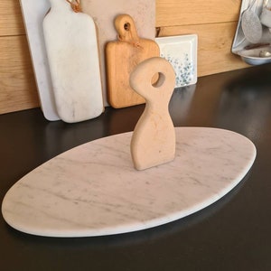 Peut inclure: Un plateau de service ovale en marbre avec une poignée en bois clair. La planche présente un motif veiné blanc et gris. Plusieurs planches à découper en bois sont à l'arrière-plan, ainsi qu'une petite assiette décorative.