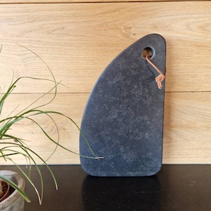 Op de afbeelding: Een zwart leisteen serveerplank met een leren lus als handvat. De plank heeft een unieke, onregelmatige vorm en een licht getextureerd oppervlak.