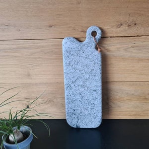Peut inclure: Une planche à découper rectangulaire longue et de couleur gris clair avec un motif moucheté. La planche a une petite poignée arrondie avec un trou pour l'accrocher. La planche est faite d'un matériau durable et est parfaite pour servir du fromage, de la charcuterie ou d'autres amuse-gueules.