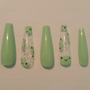 May include: Five long false nails in a green shade with a clear glitter finish. Three of the nails feature a black and white smiley face design.