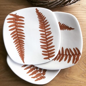 Può includere: Tre piatti in ceramica bianca con un design a felce di colore arancione ruggine. I piatti sono impilati uno sopra l'altro.