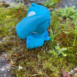 May include: A light blue figurine of a cartoon character with large ears and a white patch on its head. The figurine is sitting on a bed of green moss and small plants, with a blurred background of stone.