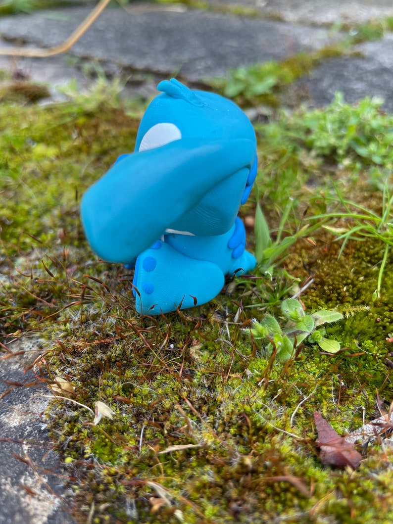 May include: A light blue figurine of a cartoon character with large ears and a white patch on its head. The figurine is sitting on a bed of green moss and small plants, with a blurred background of stone.