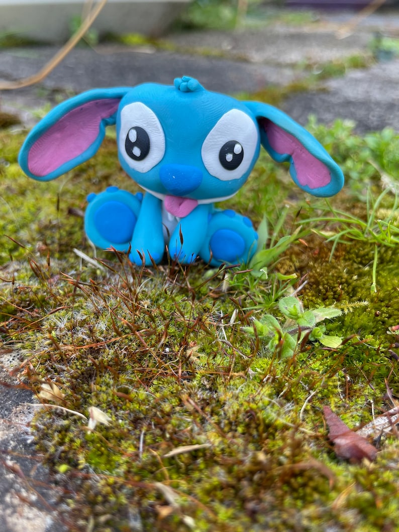 May include: A blue figurine of the character Stitch from the animated film Lilo & Stitch. The figurine has large white eyes, pink inner ears, and a pink tongue. It is sitting on a bed of green moss.
