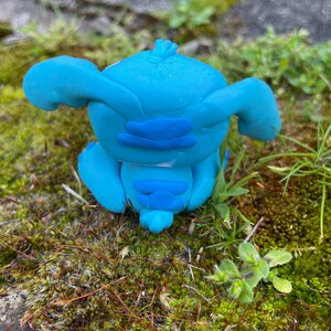May include: A handmade blue clay figurine of a cartoon character with large ears and a distinctive facial structure. The figurine is sitting on a bed of green moss and grass, with a stone surface in the background.