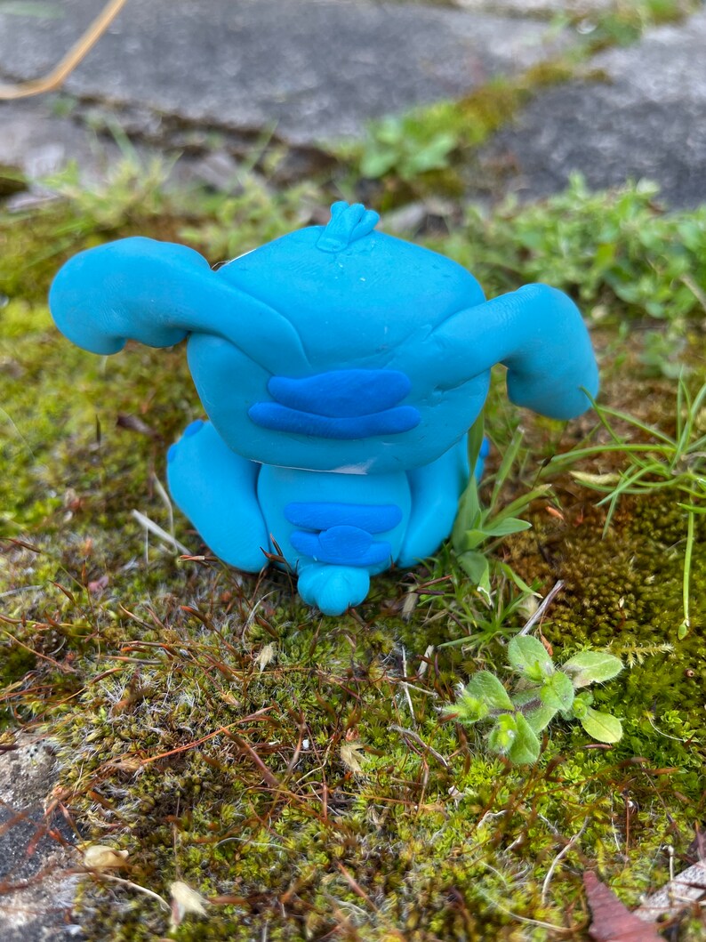 May include: A handmade blue clay figurine of a cartoon character with large ears and a distinctive facial structure. The figurine is sitting on a bed of green moss and grass, with a stone surface in the background.