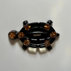 May include: Black turtle-shaped trinket with gold-colored accents. The turtle has a glossy finish and the word "HAWAII" is printed on the shell. The turtle's flippers and head are also black.