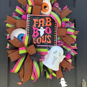 Könnte beinhalten: Ein Halloween-Kranz mit orange-schwarz gestreiften Bändern, neongrünen und rosa Bändern und dekorativen Augäpfeln. Der Kranz zeigt ein Schild mit der Aufschrift "FAB BOO LOUS" mit einer Geisterillustration. Eine weiße Geisterdekoration ist ebenfalls enthalten.