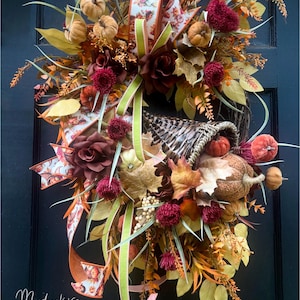 Könnte beinhalten: Ein dekorativer Kranz mit einem geflochtenen Füllhorn, gefüllt mit Miniaturkürbissen, dunkelroten Blumen und herbstfarbenem Laub. Das Arrangement enthält gemusterte Bänder und den Text Moodmakers Home Decor unten links.