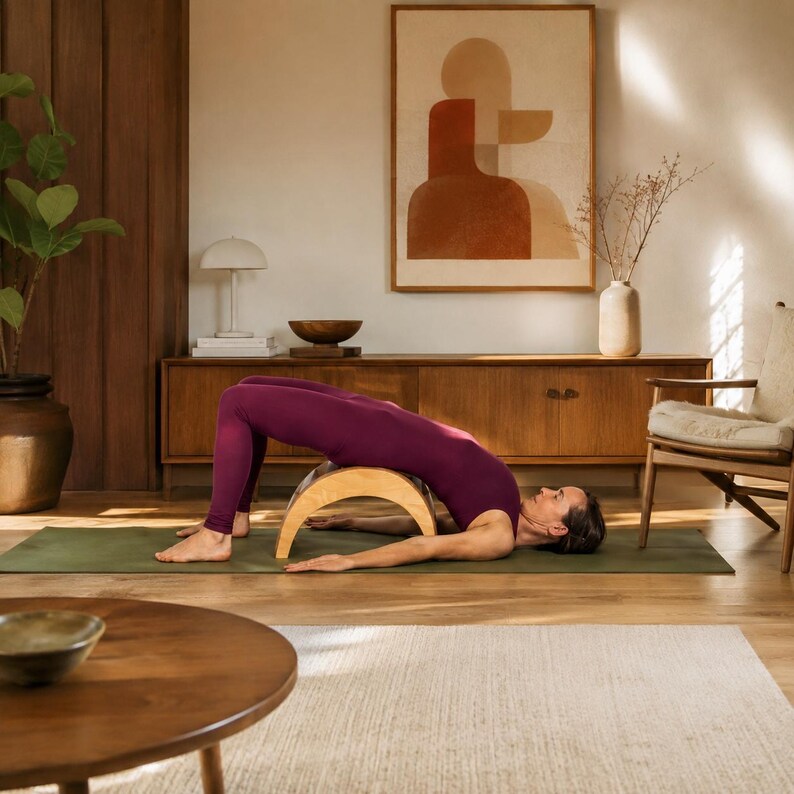 May include: A curved wooden yoga back stretcher positioned under the spine of a person wearing a dark purple bodysuit, who is lying in a bridge pose on a flat olive green exercise mat on a wooden floor in a bright, neutral-toned room.