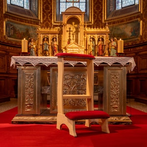 Peut inclure: Un prie-dieu en bois avec un coussin rouge, placé devant un autel d'église. L'autel est décoré d'accents dorés et présente des statues de saints. Deux bougies allumées sont visibles sur l'autel.