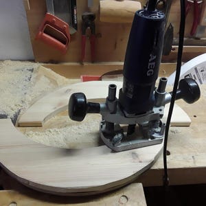 May include: A dark blue AEG plunge router with two black handles and a metal base, positioned on a curved piece of light-coloured wood. The power tool features a black cord and is used for woodworking tasks on the wooden surface.