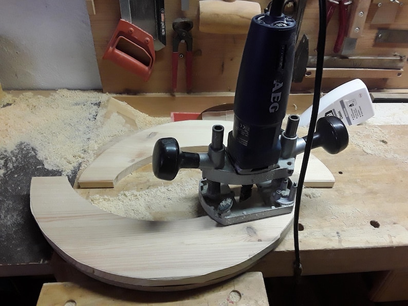 May include: A dark blue AEG plunge router with two black handles and a metal base, positioned on a curved piece of light-coloured wood. The power tool features a black cord and is used for woodworking tasks on the wooden surface.