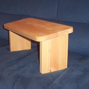 Peut inclure: Un banc de méditation en bois avec une finition naturelle. Le banc a un dessus rectangulaire et quatre pieds. Le banc est posé sur un canapé bleu.