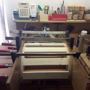 May include: A light-coloured wooden frame assembly held together by multiple metal bar clamps with red handles. The structure consists of horizontal wooden slats secured between two vertical side panels, currently undergoing a construction process.