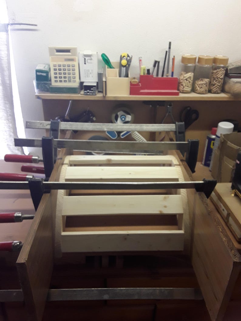 May include: A light-coloured wooden frame assembly held together by multiple metal bar clamps with red handles. The structure consists of horizontal wooden slats secured between two vertical side panels, currently undergoing a construction process.