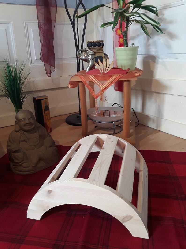 May include: A light-coloured wooden yoga back stretcher with a curved, arched design featuring four parallel slats. The structure is made of smooth, unfinished wood and is positioned on a red fabric surface in front of a seated Buddha statue.
