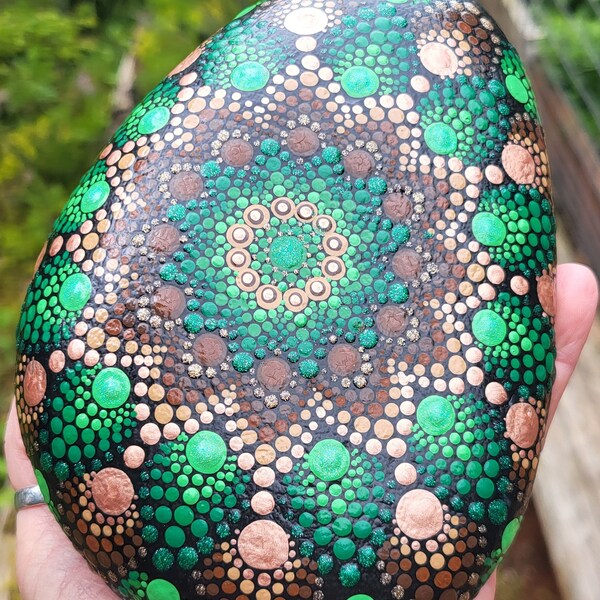 A large, oval-ish river rock painted with a beautiful green, brown & gold mandala. Has plenty of green glitter with touches of metallic gold