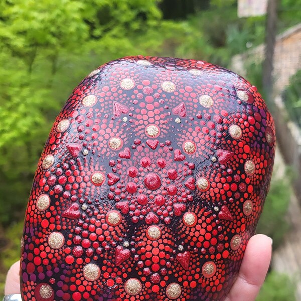 A large, irregularly-shaped river rock painted with a beautifully glittery red & gold mandala. Has some hearts painted in too!