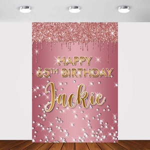 May include: Pink and gold glitter backdrop with the text "Happy 65th Birthday Jackie" in gold lettering. The backdrop is decorated with gold glitter and white diamond-shaped confetti.