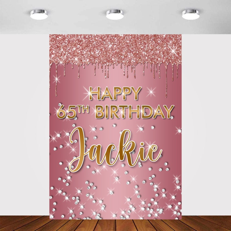 May include: Pink and gold glitter backdrop with the text "Happy 65th Birthday Jackie" in gold lettering. The backdrop is decorated with gold glitter and white diamond-shaped confetti.