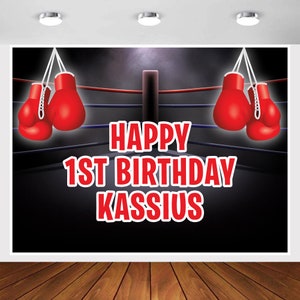 May include: A black and red boxing ring backdrop with the text "Happy 1st Birthday Kassius".  Two pairs of red boxing gloves hang from the top of the ring.