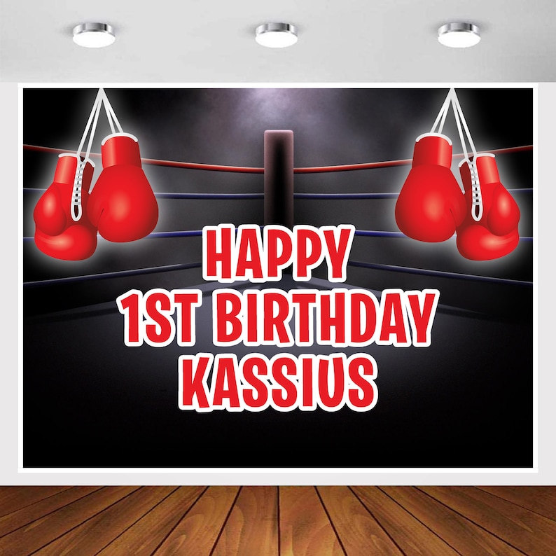 May include: A black and red boxing ring backdrop with the text "Happy 1st Birthday Kassius".  Two pairs of red boxing gloves hang from the top of the ring.