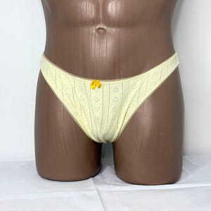 Puede incluir: Tanga amarillo pálido con un estampado de corazones y un pequeño lazo amarillo. La ropa interior tiene un delicado ribete de encaje en los bordes. La prenda se exhibe en un maniquí.